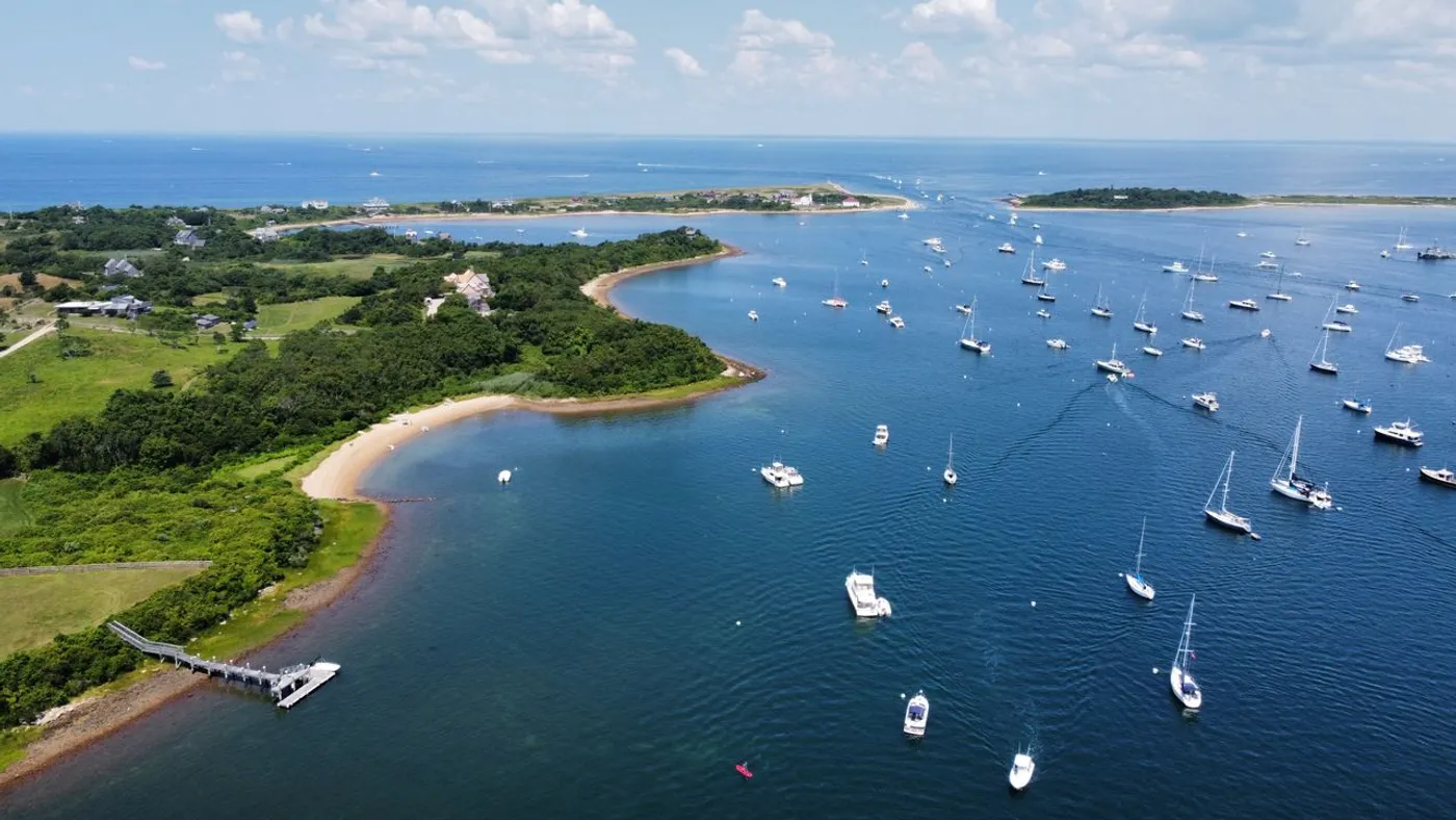 Aerial view of the Great Salt Pond at New Harbor: dozens of sailboats moored in blue water inside the island's protected pond.