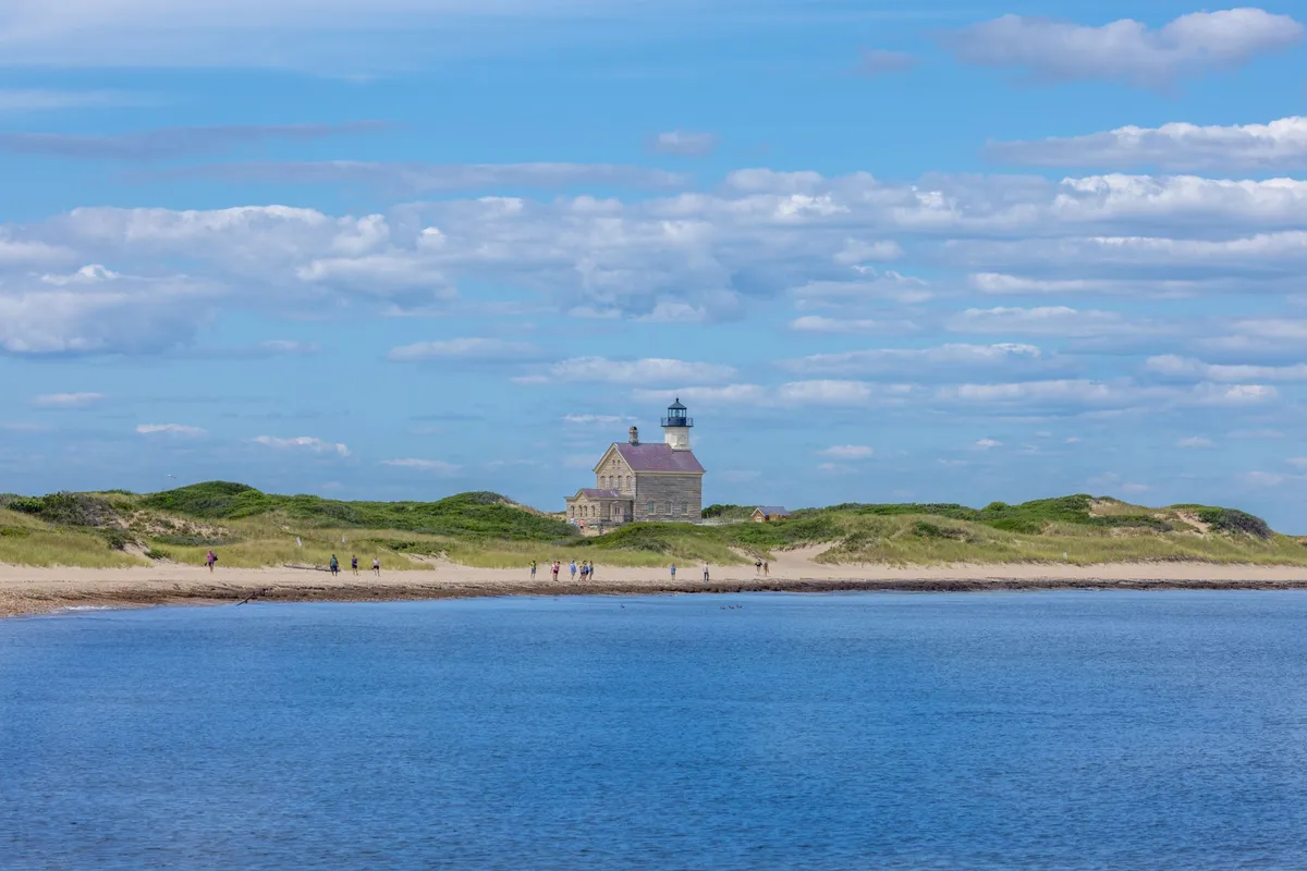 The granite North Lighthouse at Sandy Point, seen from the water with walkers on the beach and dunes behind.