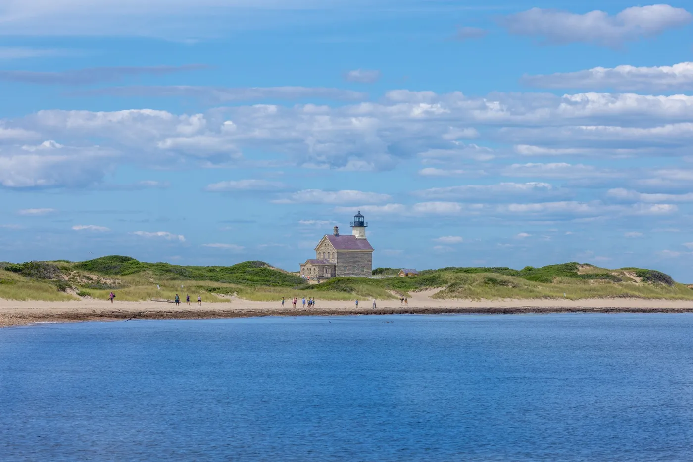 The granite North Lighthouse at Sandy Point, seen from the water with walkers on the beach and dunes behind.
